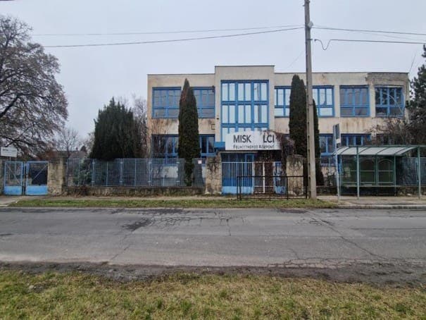 Háromszintes, kék nyílászárós intézményi épület kerítéssel